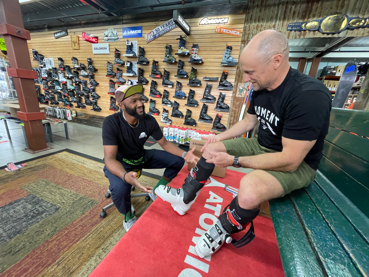 Staff members assists customer with a ski boot fitting while customer is seated on a green bench. Ski boots are visible on the wall behind.