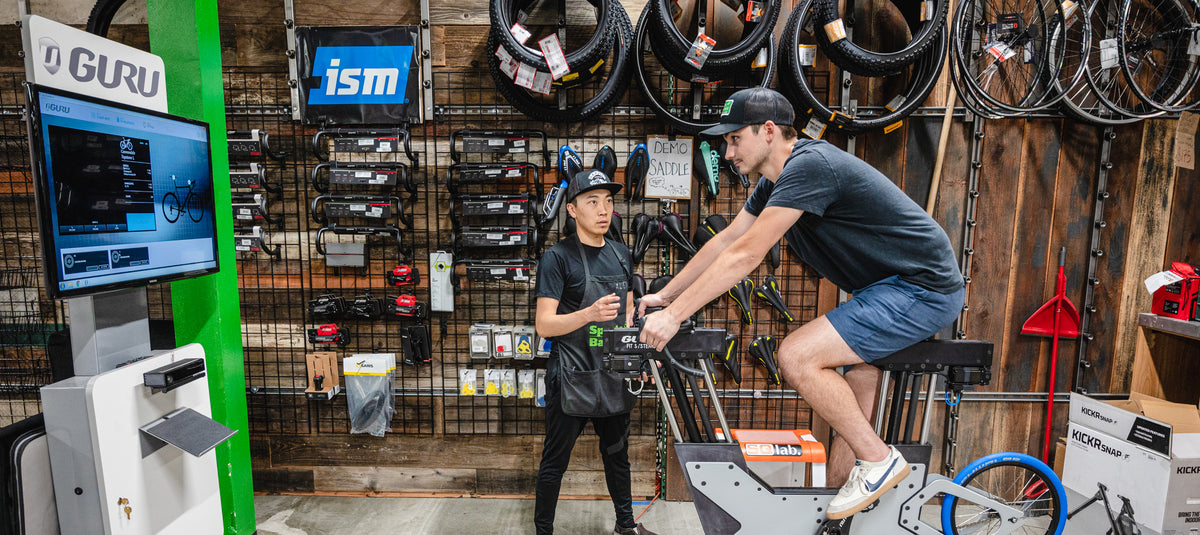 Staff member helps a customer with a bike fitting on a futuristic looking GURU bike fitting machine, in front of a wooden wall filled with bike components.