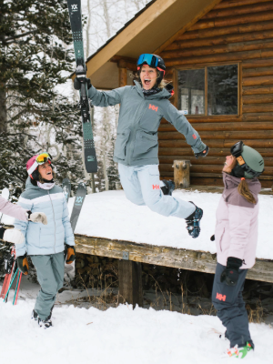 Three people in ski gear celebrate on a snowy porch in front of a wooden cabin. The central person jumps joyfully while holding skis, wearing a gray ski jacket and light pants, with two others in light blue and pink jackets nearby.